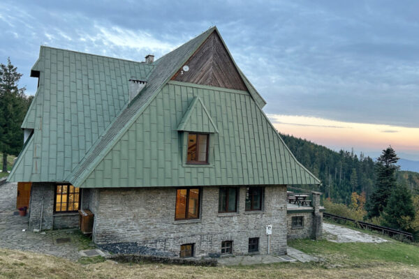 Zdjęcie schroniska PTTK na Przechybie. Jasny szary kamień elewacji i duży spadzisty jasno zielony dach. W tle panorama gór.